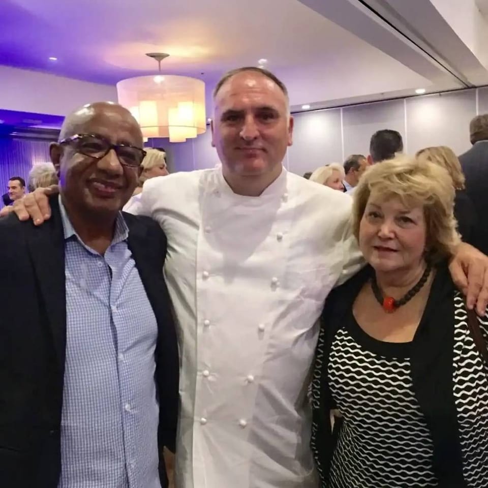 Chef in a white jacket poses with two guests at a lively restaurant event under purple lighting.