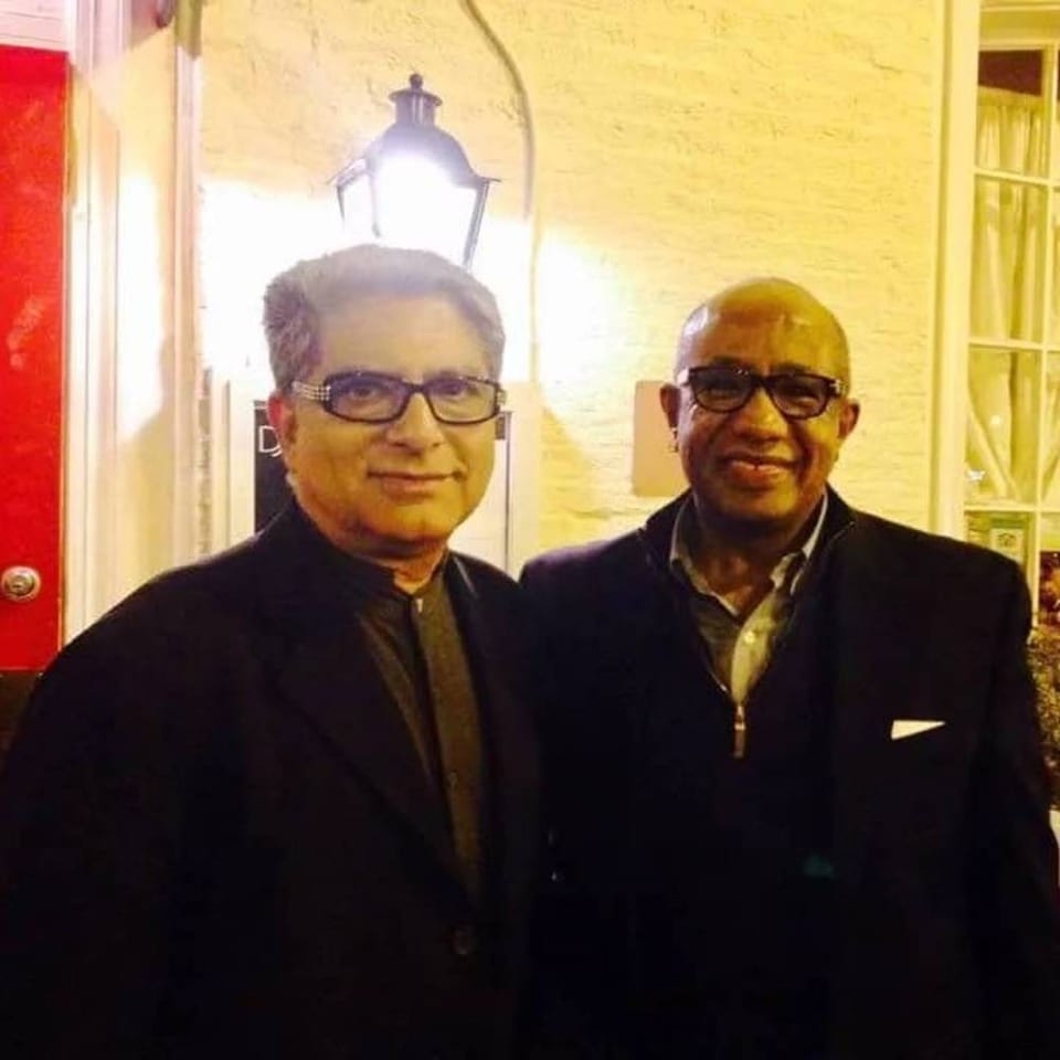 Two men in dark jackets and glasses pose beneath a glowing lantern outside a warmly lit building.