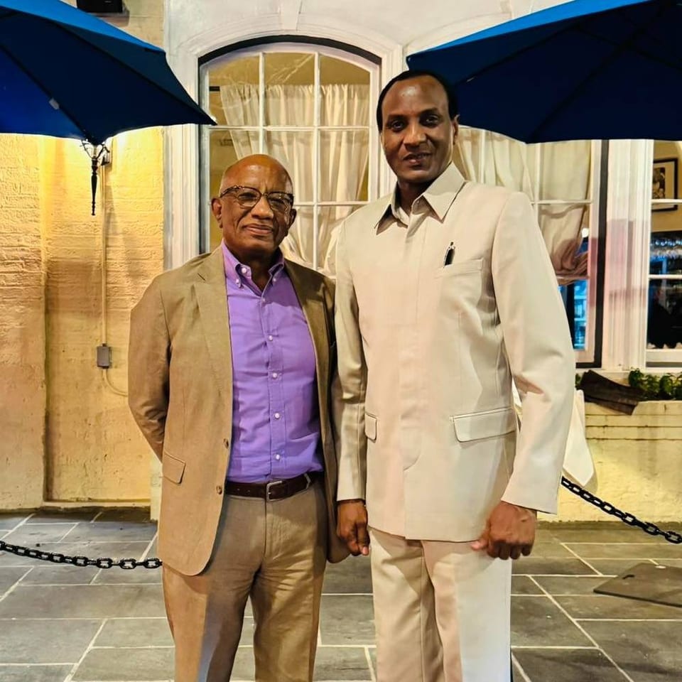 Two men pose outside a restaurant beneath blue umbrellas, with a warmly lit window and stone-paved courtyard behind them.