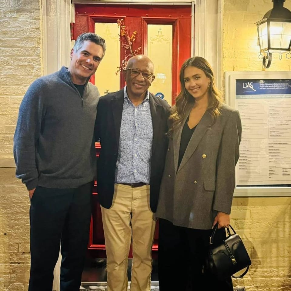 Three smiling adults pose outside a restaurant’s red door beside a framed menu and wall lantern.