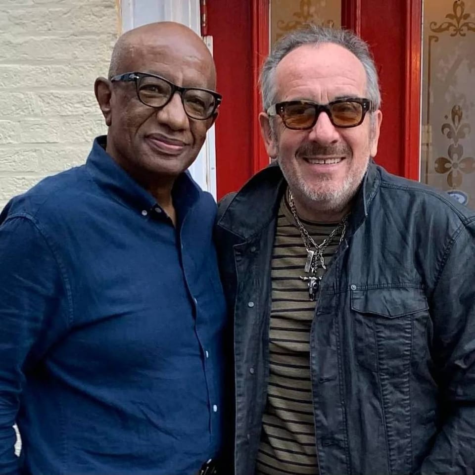 Two smiling men wearing glasses pose together outside a red-painted doorway.
