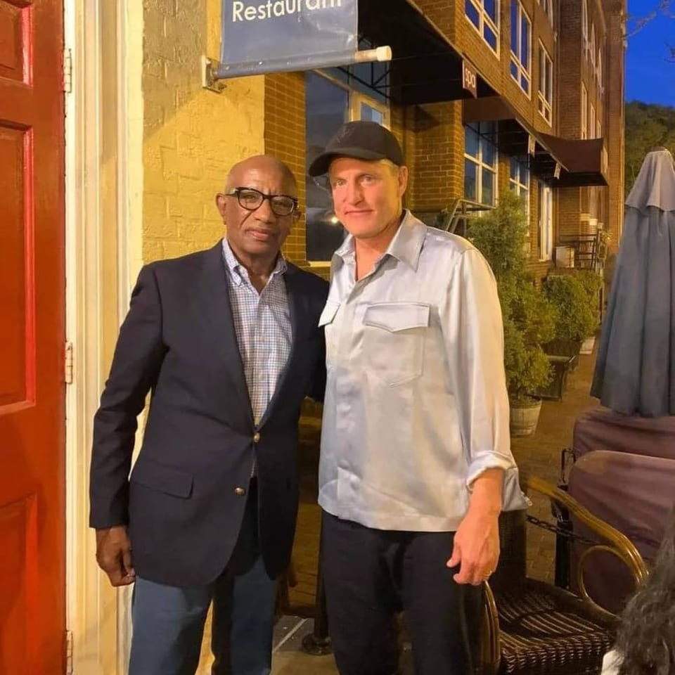 Two men pose outside a restaurant entrance beside outdoor seating and a covered patio umbrella.