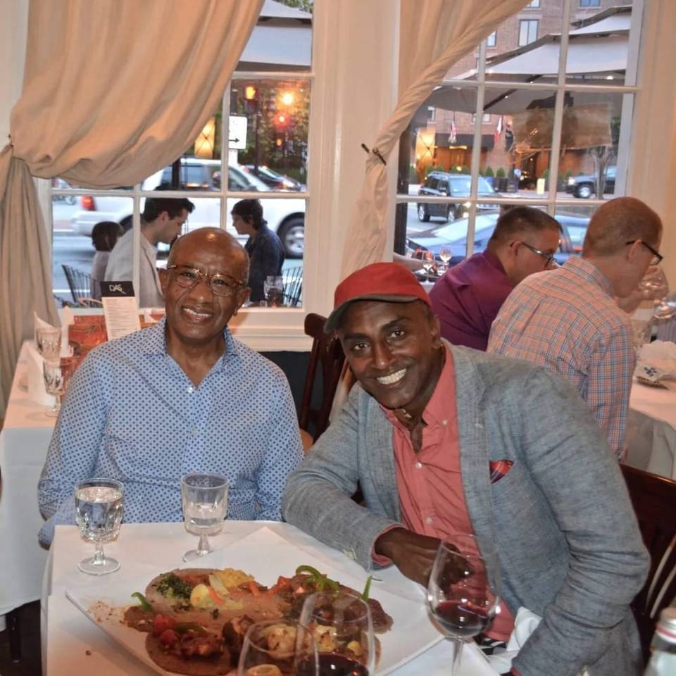 Two smiling diners pose beside a plated meal and wine at a warmly lit restaurant table.