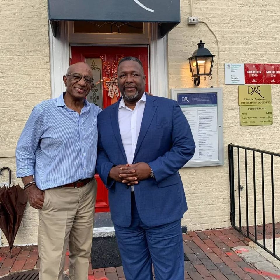 Two men pose outside DAS Ethiopian Restaurant beneath Michelin recognition plaques and a wall-mounted lantern.