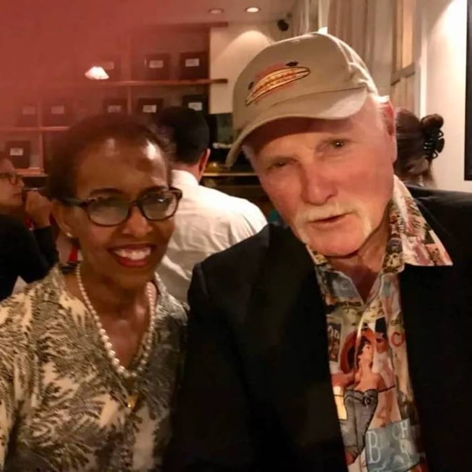 Two smiling people pose together inside a lively restaurant, with diners and wooden shelving visible in the background.